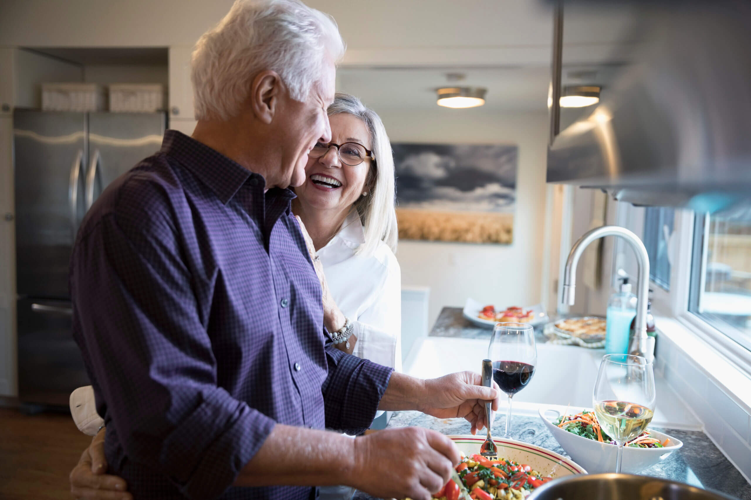 Affectionate senior couple laughing cooking and drinking wine in kitchen