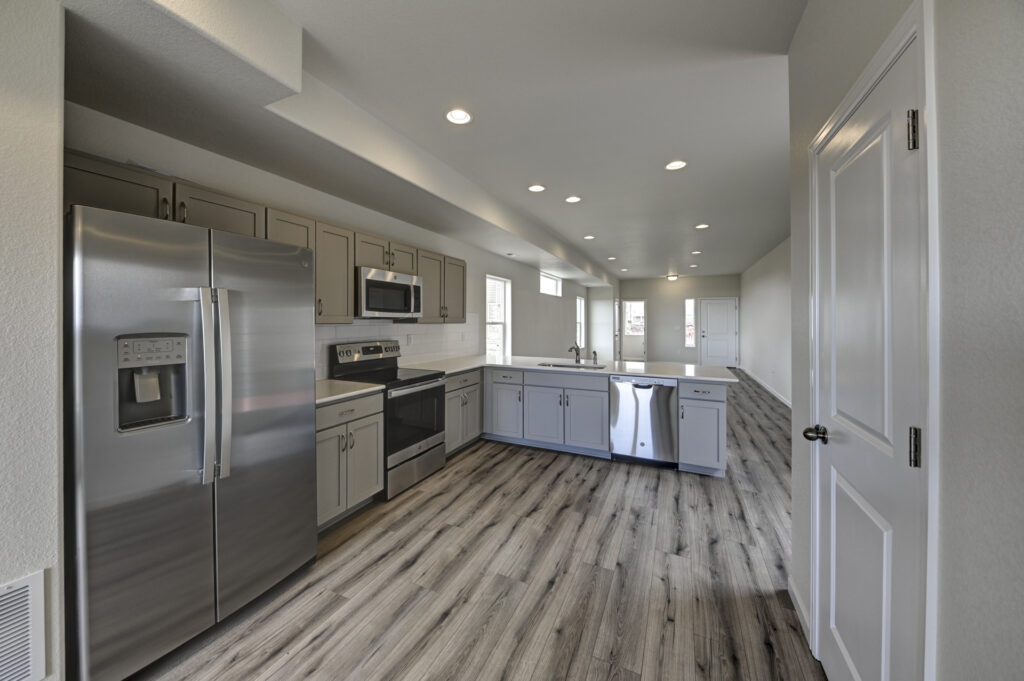 Open kitchen with stainless appliances, gray cabinets and wood-look vinyl flooring under recessed lights.