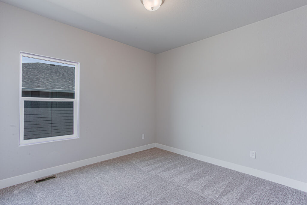 Empty carpeted room with single window and light-gray walls, simple ceiling fixture.