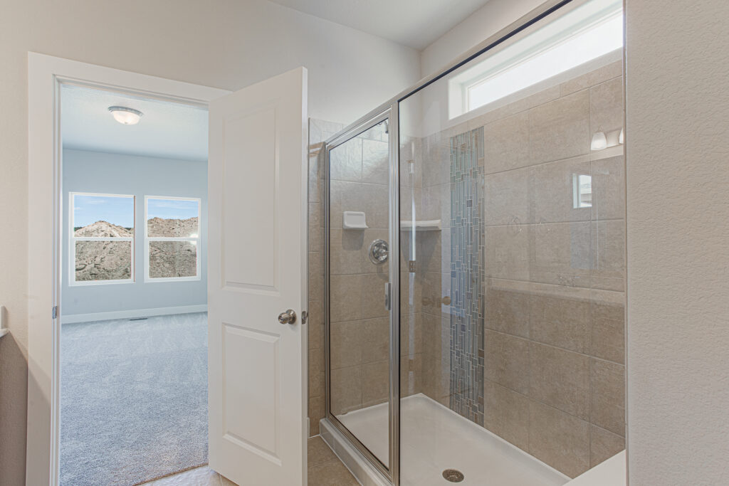 Glass-enclosed tiled shower next to an open door showing a carpeted room with three windows.