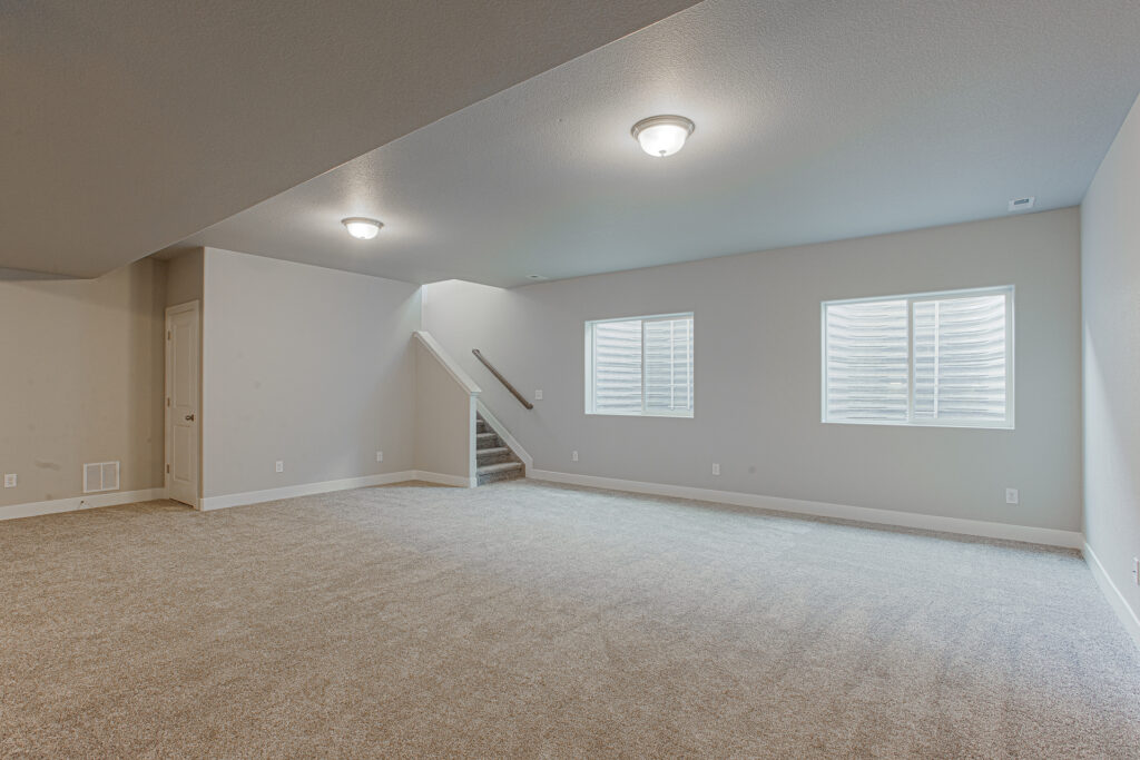 Open finished basement room with two window wells and carpeted stairs to the right.