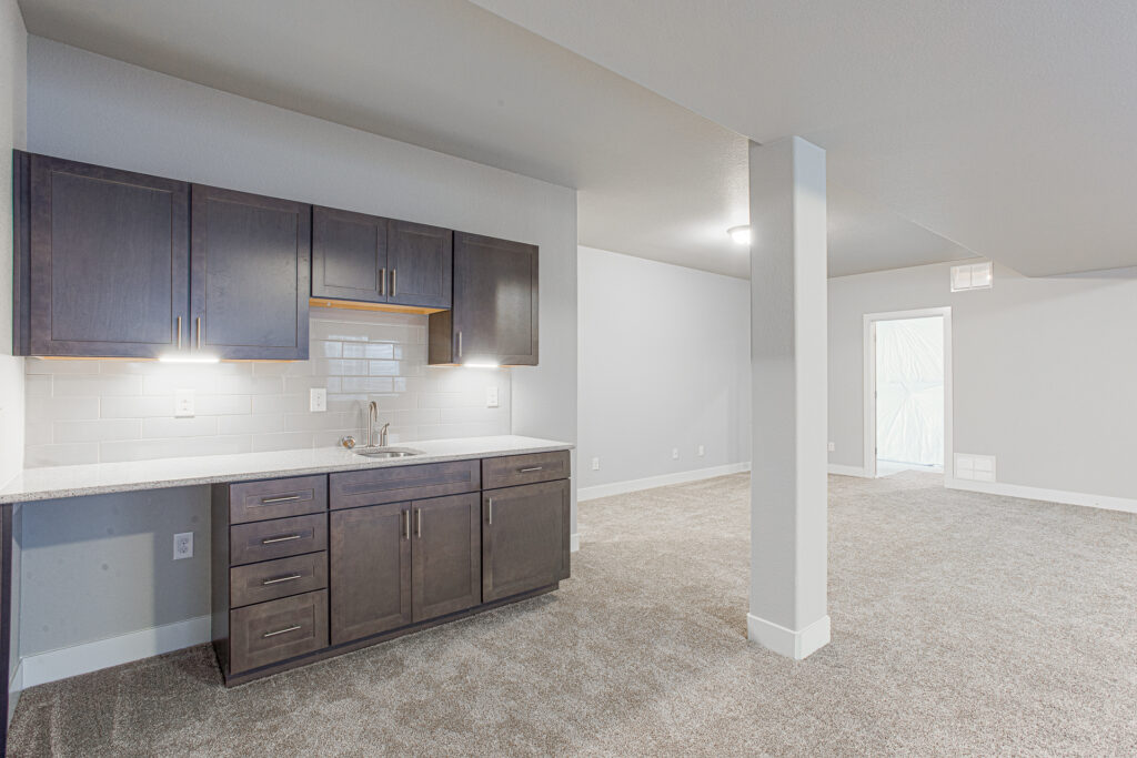Basement kitchenette with dark wood cabinets, under-cabinet lighting, and carpeted open area.