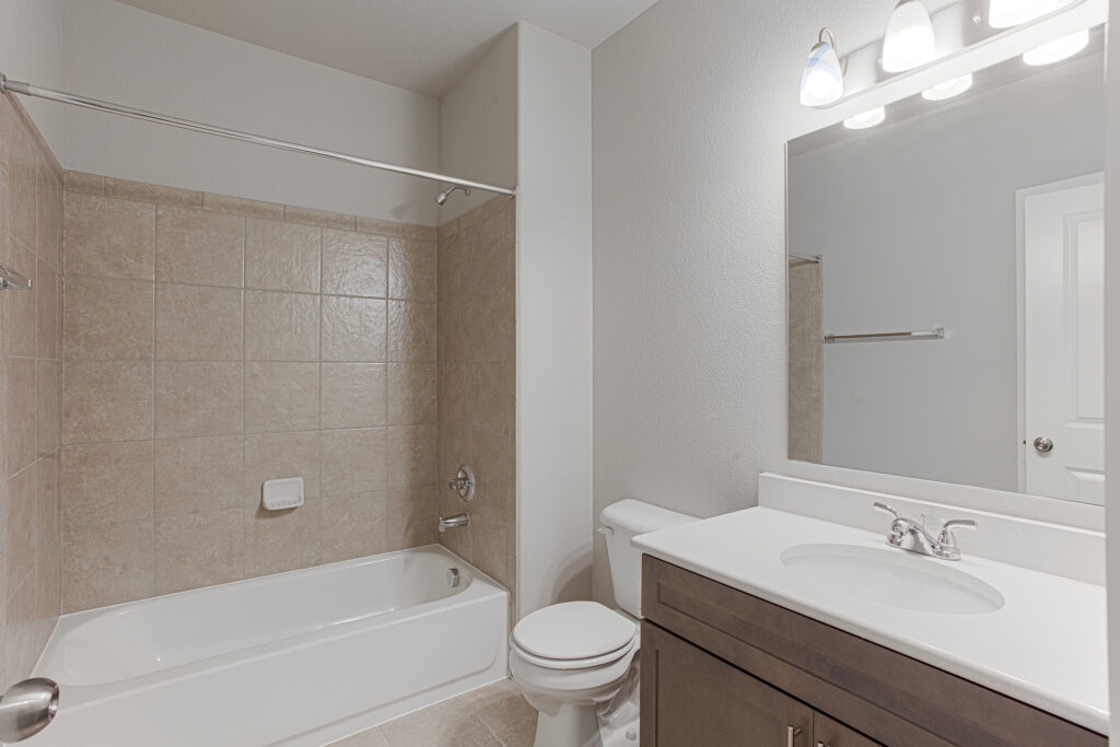 Bathroom with tiled tub/shower surround, white tub, toilet, and vanity with mirror and lights.