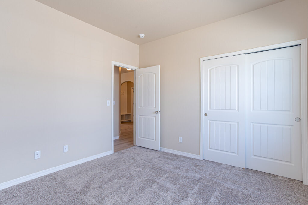Empty bedroom with beige walls, carpeted floor, white bi‑fold closet doors and an open doorway.