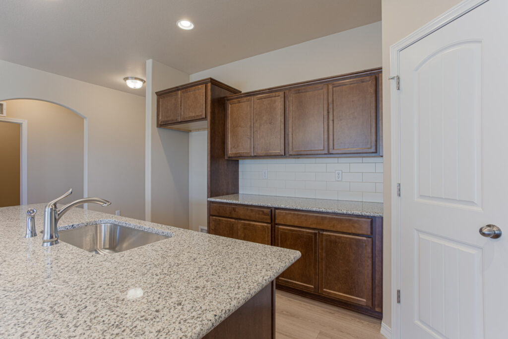 Kitchen island with sink and granite countertop facing dark wood cabinets and subway tile backsplash.