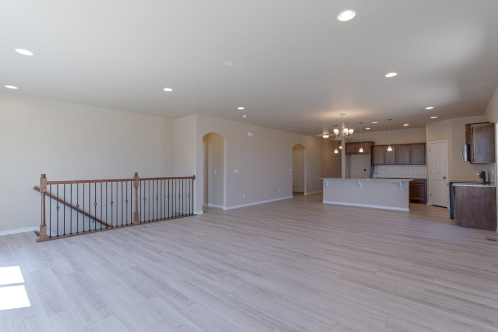 Large open living area with light wood floors, neutral walls, and staircase railing to left.