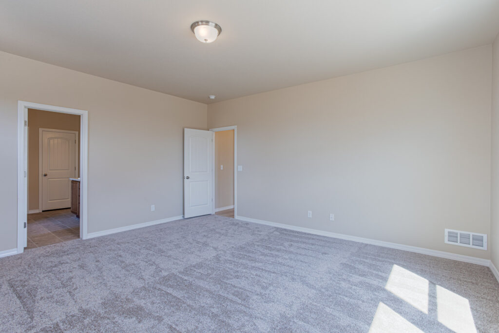 Empty carpeted bedroom with neutral walls, ceiling light, and two doors leading to adjoining spaces.