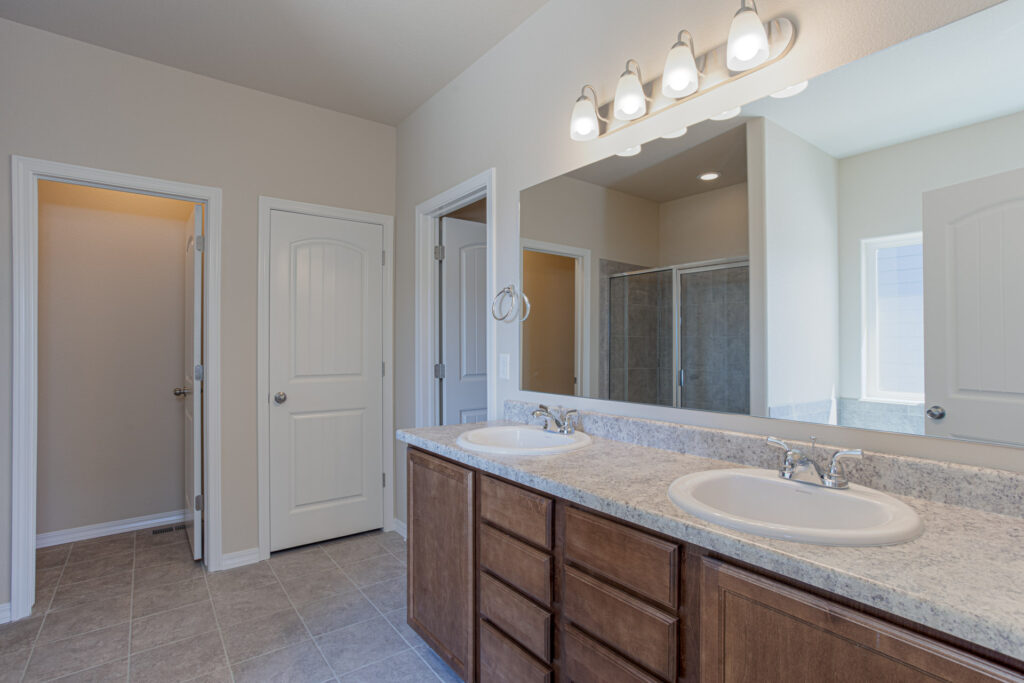 Double-sink vanity with large mirror, overhead lights, and two closed doors visible in the background.