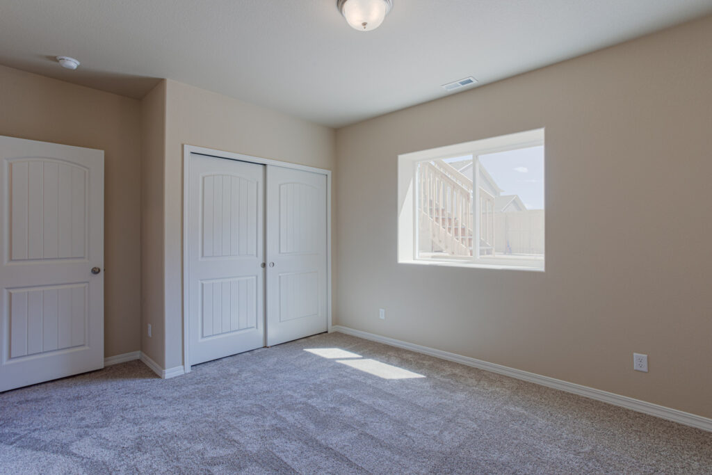 Empty carpeted bedroom with closet and a rectangular window looking out to backyard stairs.