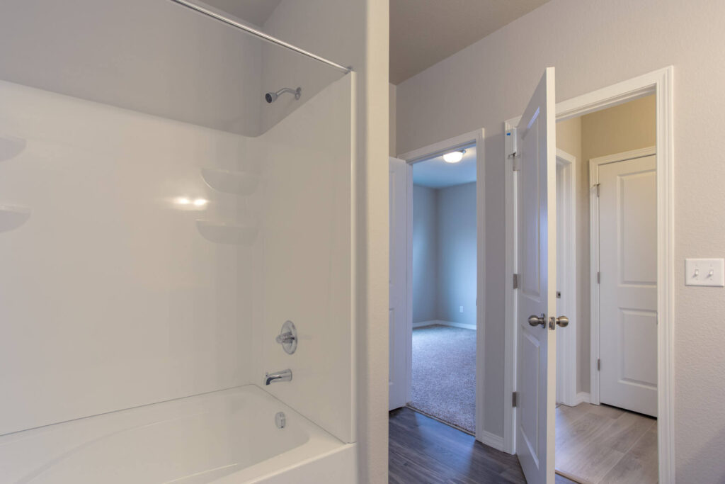 View of a white bathtub and shower next to an open doorway leading to a carpeted room.