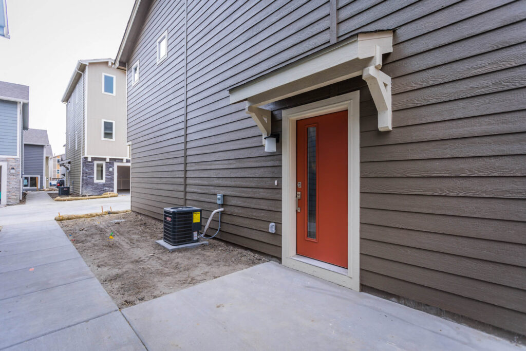 Side entrance of a house with a red front door, brown horizontal siding, and an exterior HVAC unit.