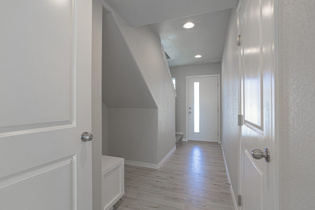 Narrow entry hallway with light wood-look flooring, white walls, and a front door with vertical glass panel.