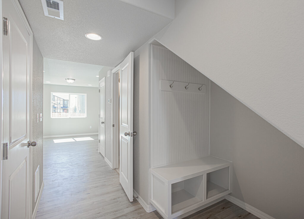 Hallway with under-stair storage bench and wall hooks, light floors, and sunlight from a distant window.