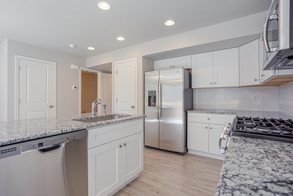 Angle view of kitchen island, sink, fridge, and gas stove with consistent white cabinetry and light floors.