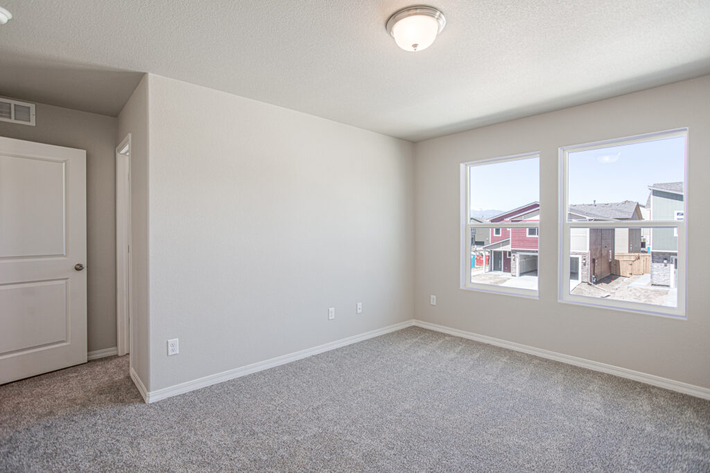 Empty carpeted bedroom with two windows overlooking neighboring houses and neutral walls.