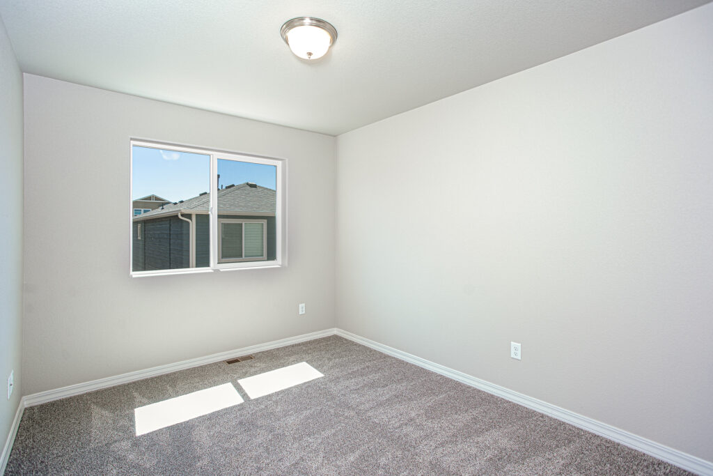 Small empty room with single window, neutral walls, and gray carpet illuminated by daylight.