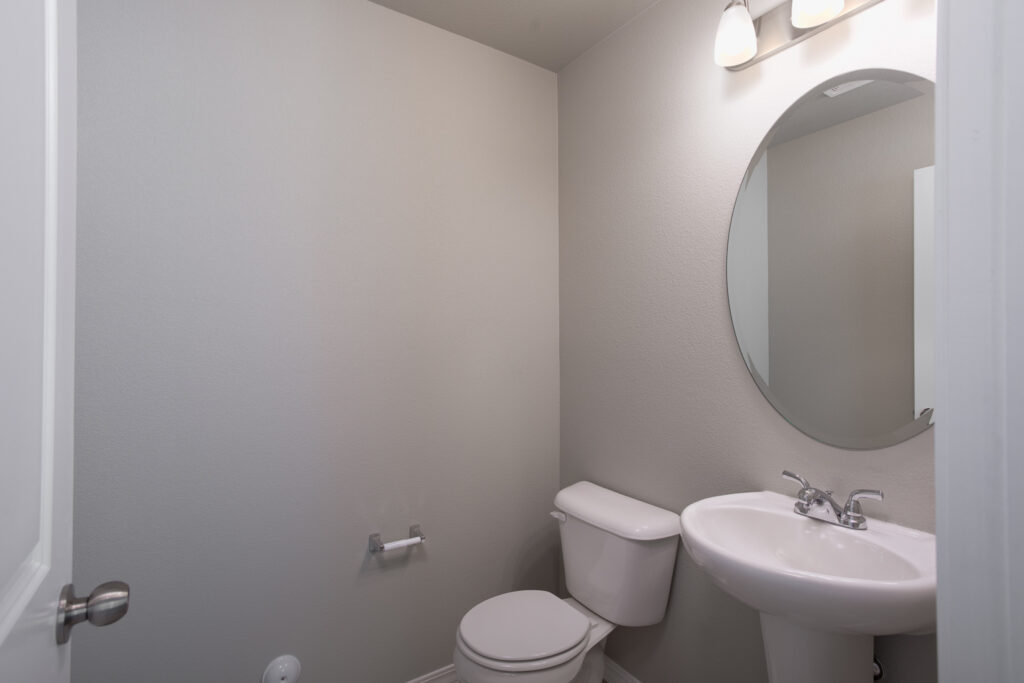 Small half-bath with pedestal sink, round mirror and toilet under soft lighting.