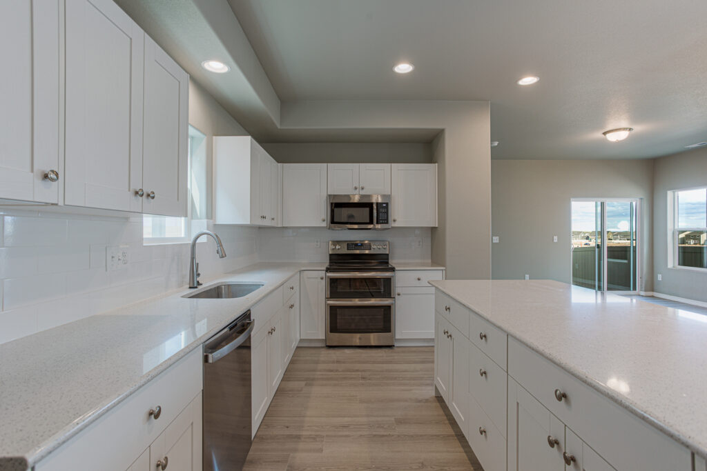 Perspective view of a white kitchen with island, dishwasher, double oven range, and open living area beyond.