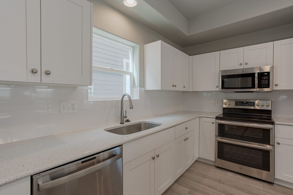 Kitchen sink wall with window, white cabinets, and stainless steel double oven and dishwasher.