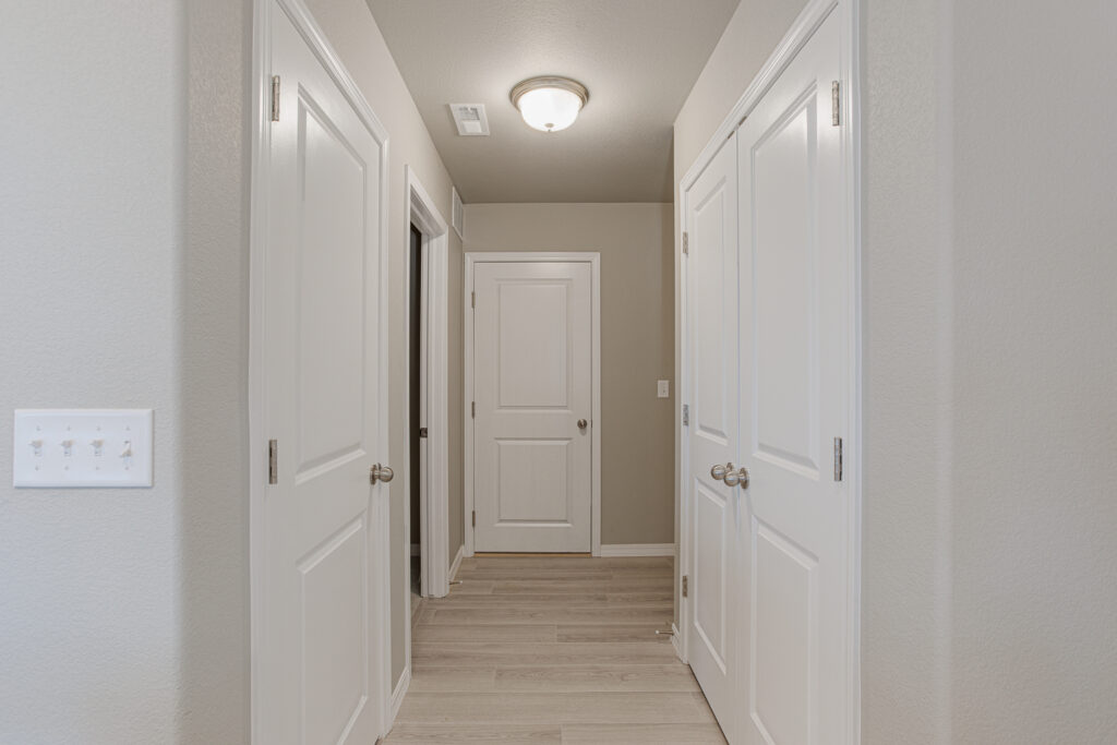 Interior hallway with multiple closed white doors and wood-look flooring under a ceiling light.