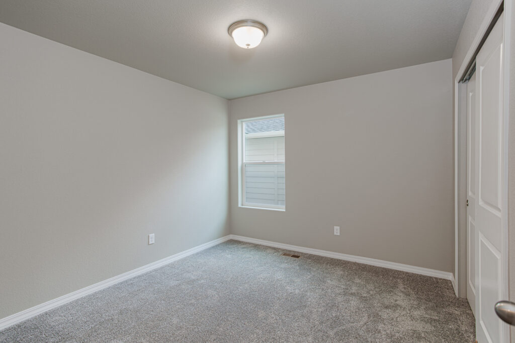 Empty small bedroom with a single window, grey carpet and neutral painted walls.