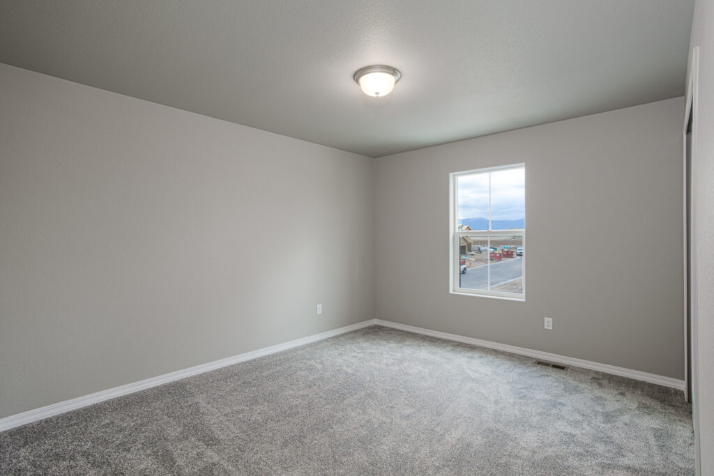 Empty carpeted room with a single window and neutral gray walls under a flush ceiling light.