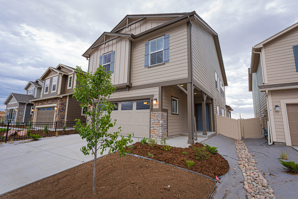 Two-story beige suburban house with attached garage, small front tree and mulch bed.