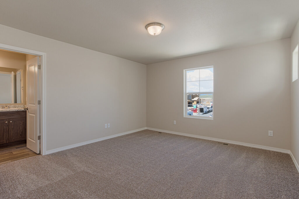 Empty carpeted bedroom with single window and neutral walls, view into an adjacent bathroom.