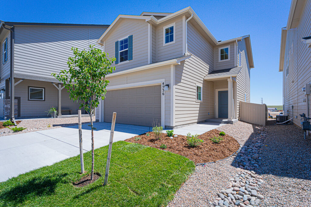 Angled view of a two-story beige house with planted beds, a young tree, and a side gravel pathway.