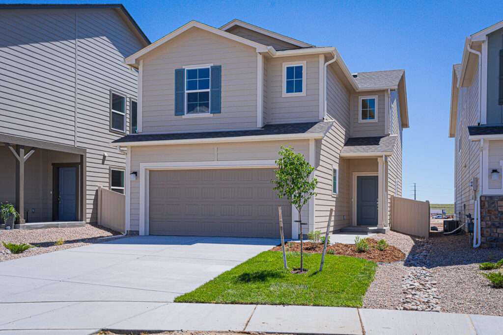 Two-story beige house with double garage, small front lawn and concrete driveway under clear sky.