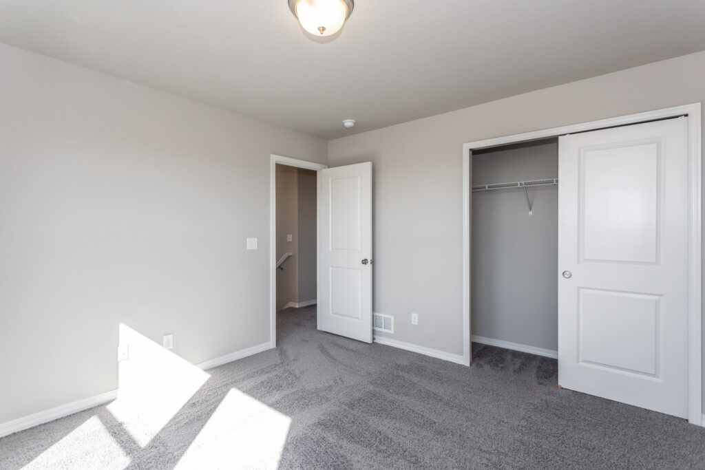 Sunlit empty bedroom with open door and closet, gray carpet and beige walls.