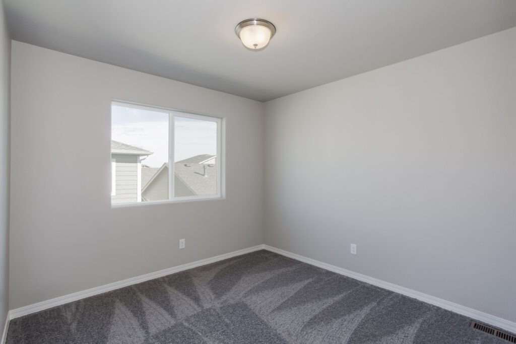 Small empty room with single window, gray carpet and plain neutral walls.