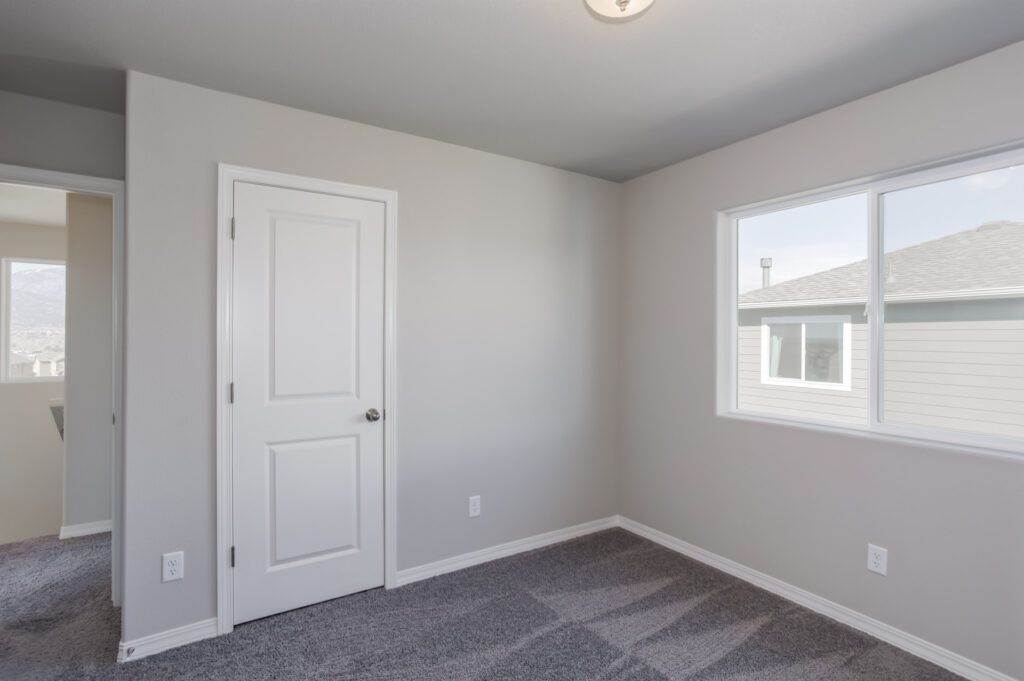 Empty small bedroom with gray carpet, light gray walls, white door and a window showing neighboring roof.
