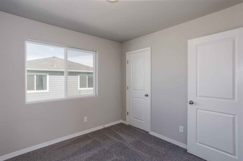 Small empty bedroom with large window, closet door and gray carpeting.