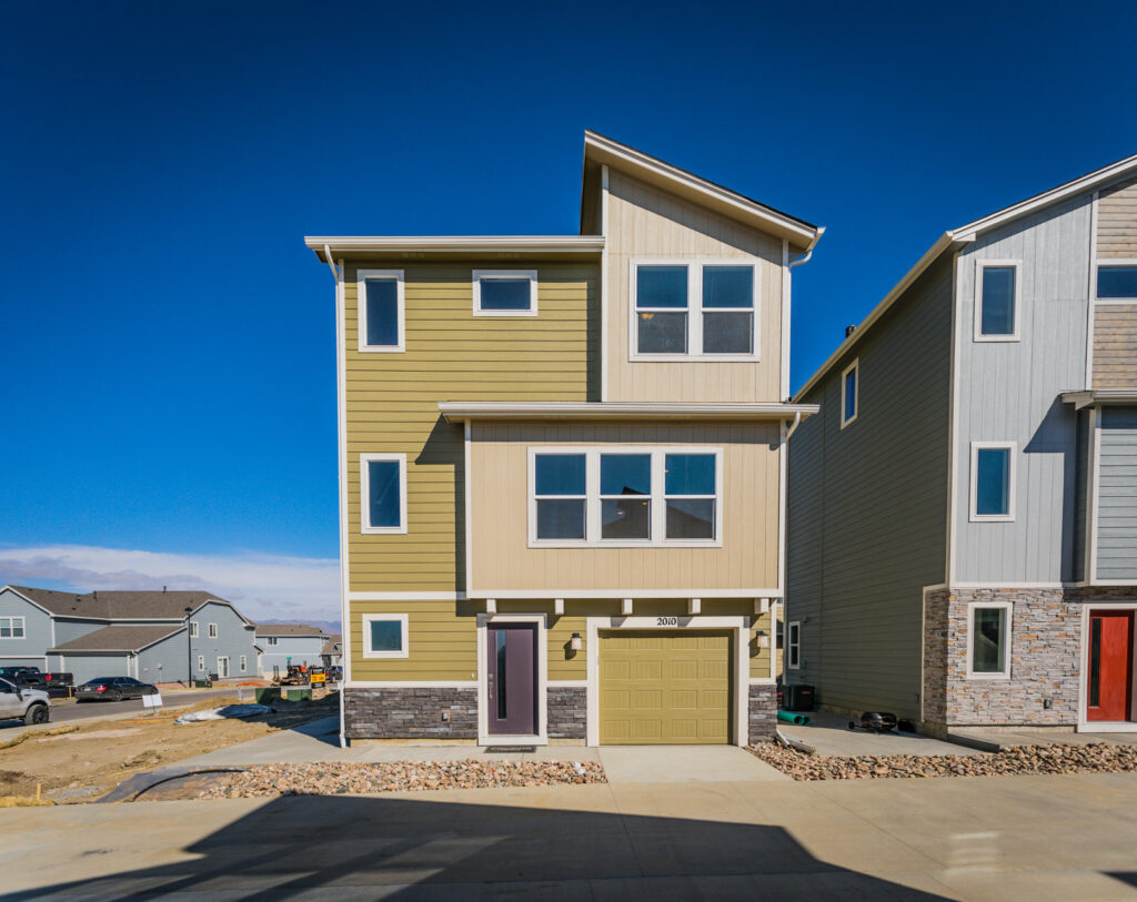 Three-story modern townhouse with green and beige siding, single-car garage, and stone accent base.