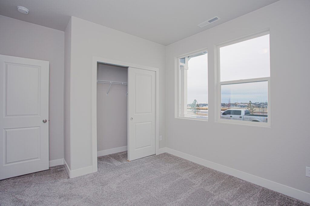 Small empty room with open closet, two windows, and gray carpet under soft natural light.