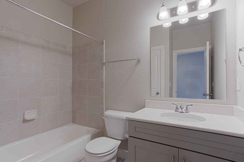 Bathroom with bathtub and tiled surround, toilet, vanity with sink, and illuminated mirror light fixture.