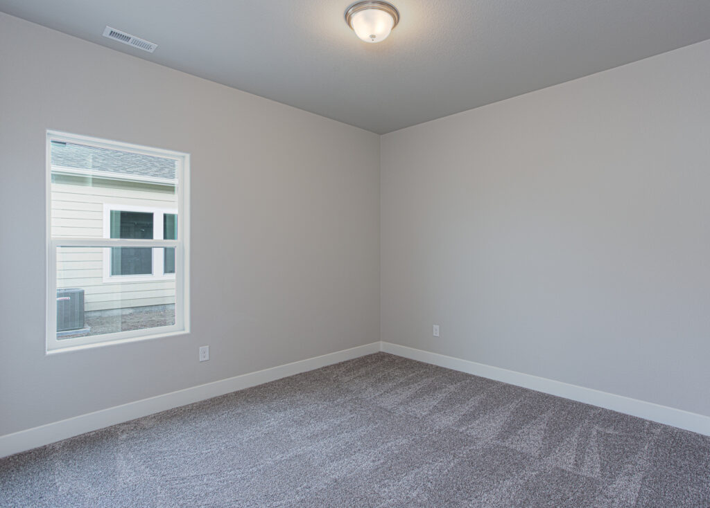 Empty small bedroom with gray carpet, single window, neutral gray walls and ceiling light.