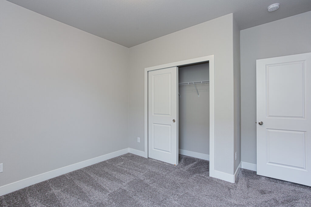 Empty bedroom showing open sliding closet door, gray carpet and neutral painted walls.