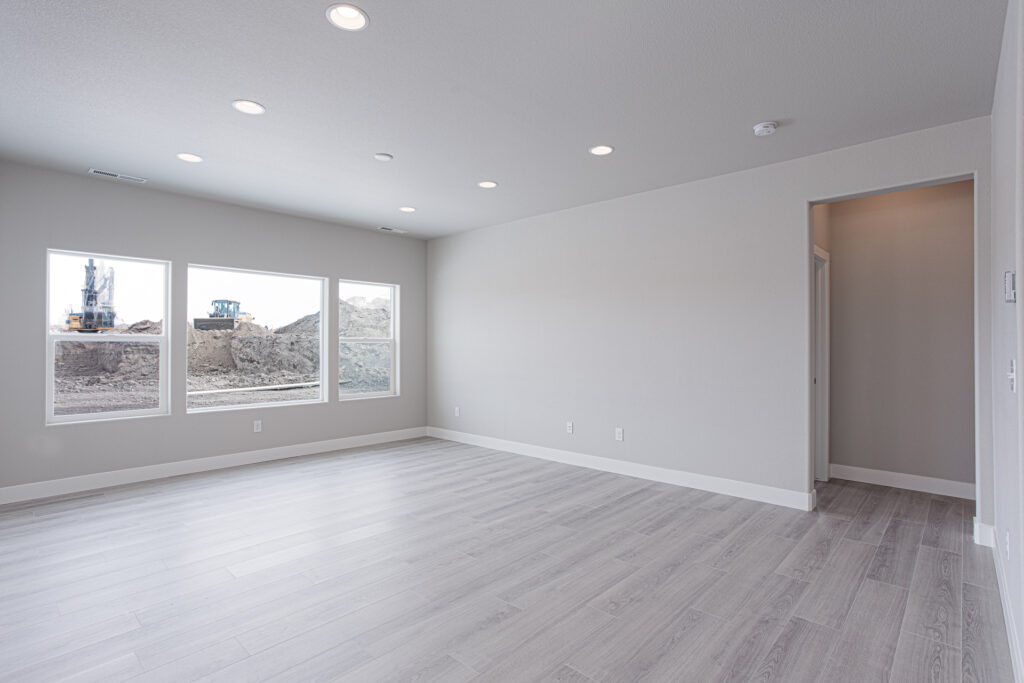 Large empty living room with three windows looking out to a construction site and recessed ceiling lights.