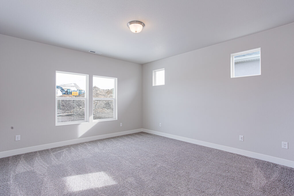 Empty carpeted bedroom with two large windows and two small high windows, ceiling light fixture.
