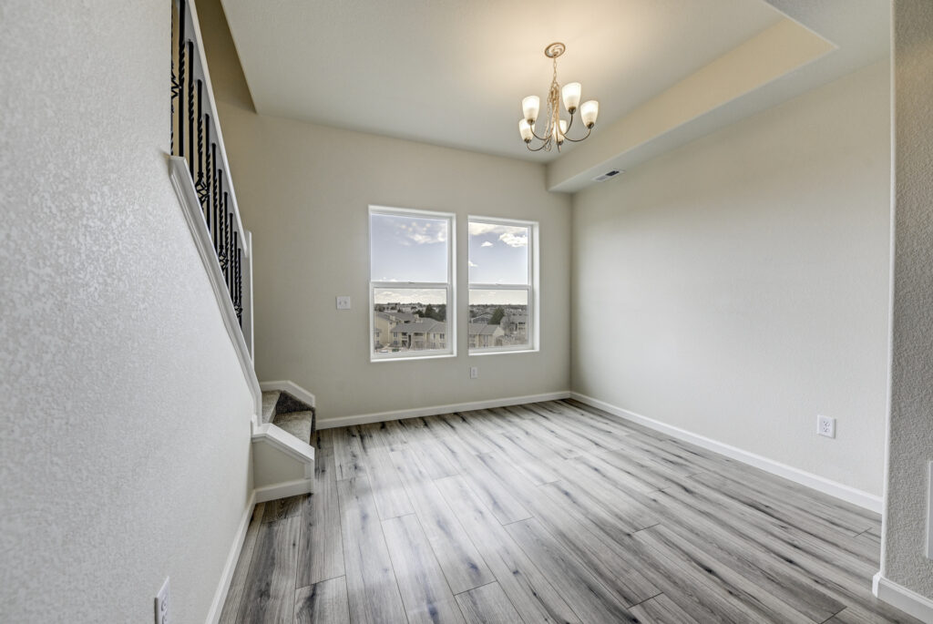 Empty dining or sitting area with two windows, chandelier and gray wood-look flooring near a staircase.