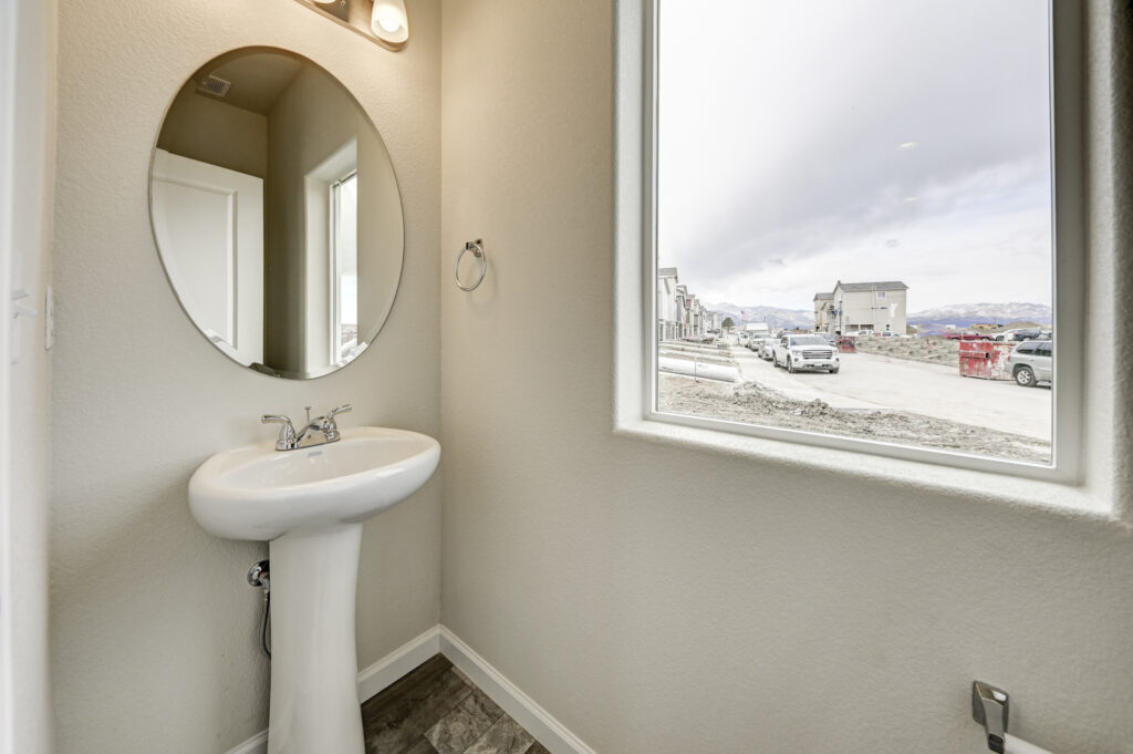 Small powder room with pedestal sink, round mirror and a window overlooking a street under construction.