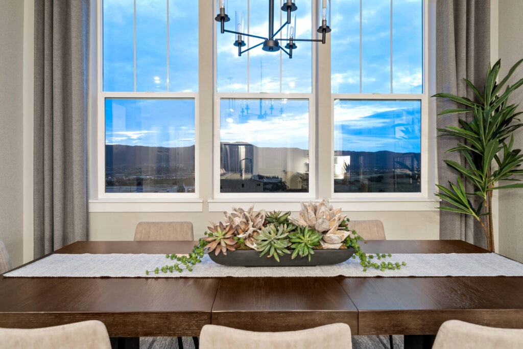 Dining table with succulent centerpiece in front of three large windows showing evening sky and distant hills.