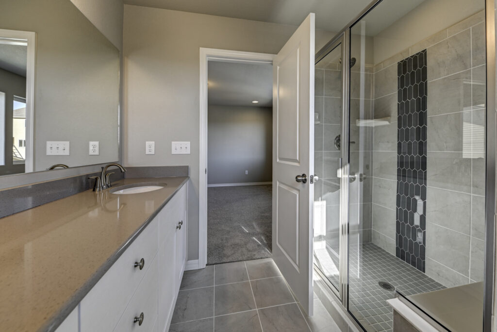 Bathroom view toward open door, showing vanity on left and glass-enclosed tiled shower on right.