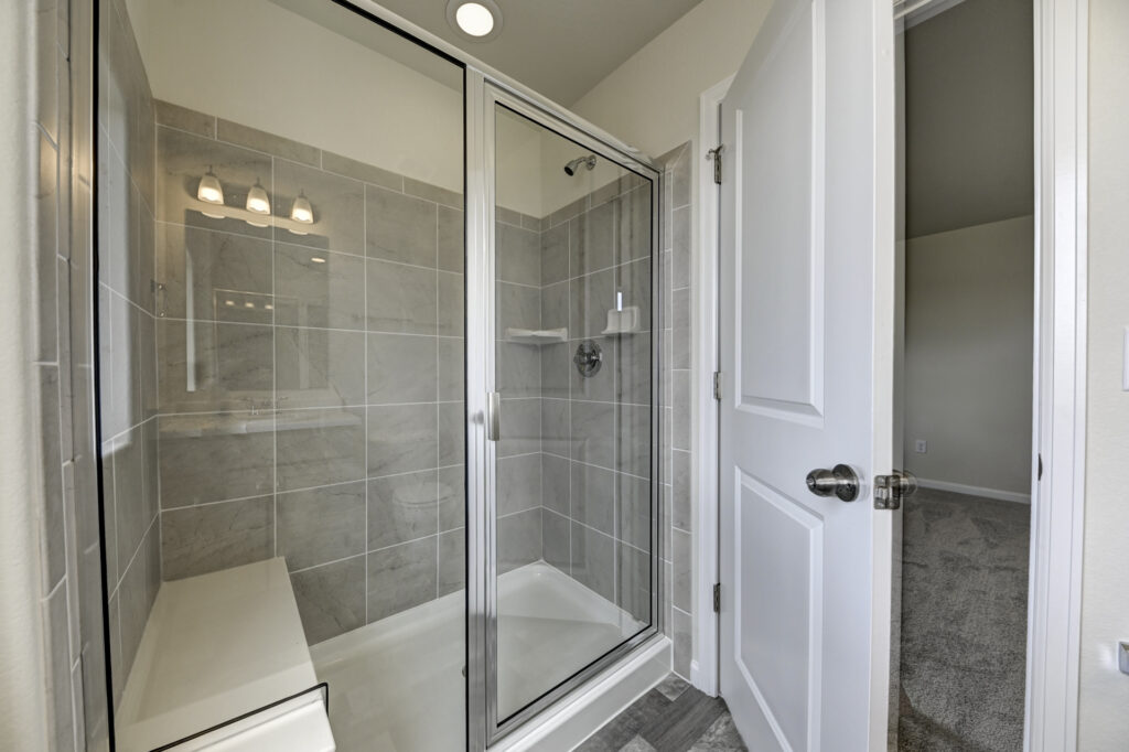 Tiled walk-in shower with glass sliding door and bench, neutral gray tiles and chrome fixtures.