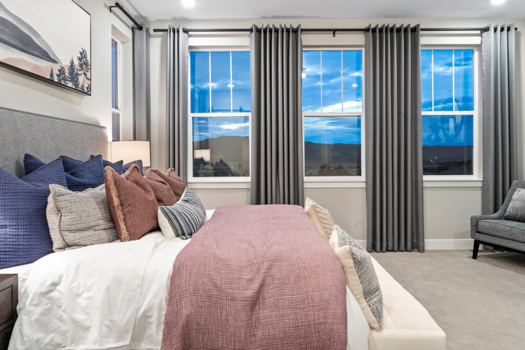 Staged bedroom with bed facing three large windows and gray curtains at dusk.