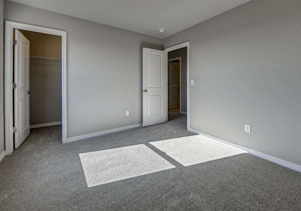 Empty carpeted bedroom with open closet and two doors, sunlight patch on floor.