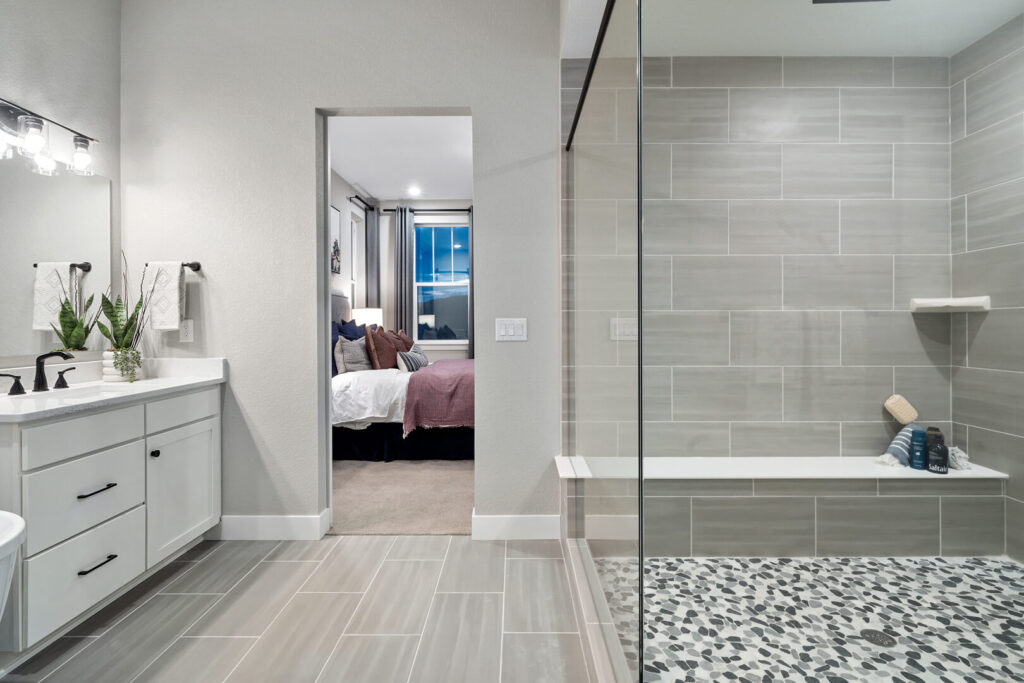 Modern bathroom with tiled walk-in shower, white vanity, and view into a carpeted bedroom.
