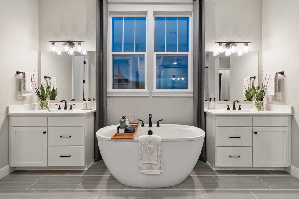 Freestanding bathtub centered between two vanities beneath a double window with curtains.
